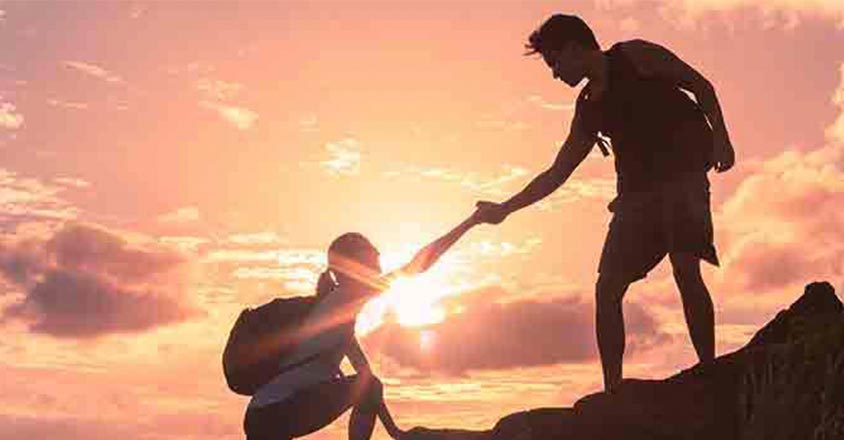 person helping another climb cliff with sunset in background