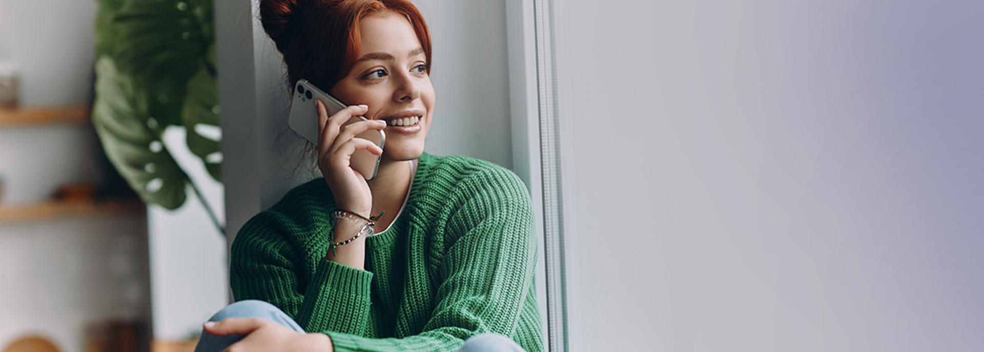 A smiling young woman with red hair pulled into a bun sits by a window and talks on her cell phone.