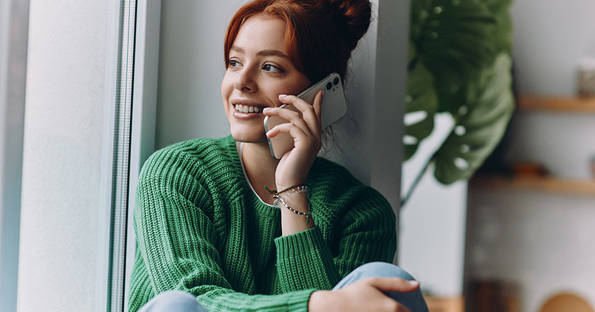 A smiling young woman with red hair pulled into a bun sits by a window and talks on her cell phone.