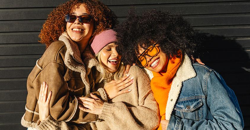 three young, diverse women hugging and smiling outside wearing jackets and sun shining