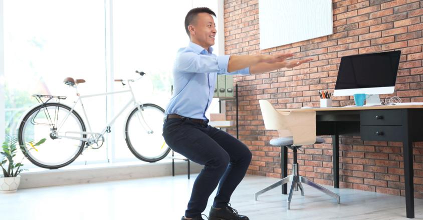 In a bright, naturally lit office, an adult man with short dark hair wearing a light blue button-down shirt, dark pants, and black shoes performs squats in front of a desk, computer, and brick wall. A bicycle stands near a window. The setting and subject suggests the concepts of active workplace, balance, and healthy habits. 