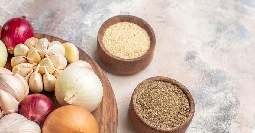 A wooden board holds a collection of culinary delights: red and white onions, fragrant garlic bulbs, and two wooden bowls filled with spices. The photo is shot in bright daylight, with a focus on the natural textures and colors of the ingredients, creating an inviting and wholesome mood. 