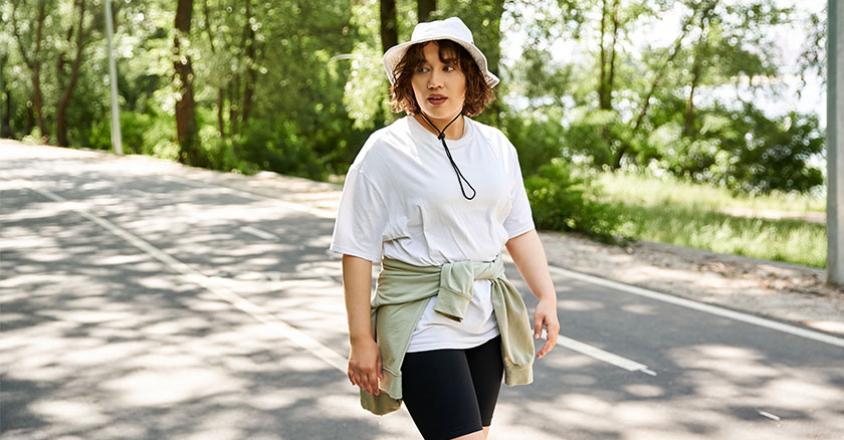  A woman with wavy dark hair walks on a paved path flanked by a verdant landscape. She is wearing a white t-shirt and black shorts, with a green sweater tied around her waist and a white hat. The background shows trees and green vegetation.    