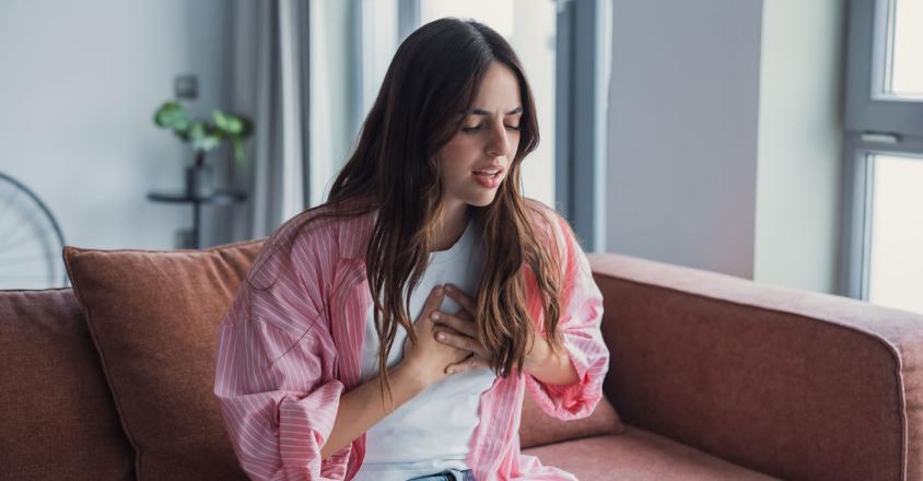 A young woman with wavy brown hair sits on a couch in a home interior. She wears a pink striped shirt, white t-shirt, and light blue jeans. She clutches her chest with both hands and appears to be in distress. The scene is brightly lit by daylight streaming in from nearby windows