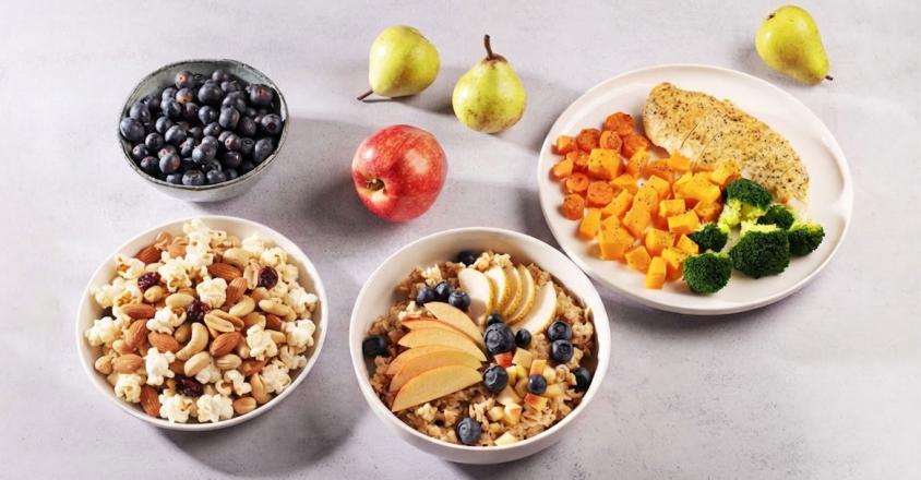 Eye-level shot presents a variety of healthy food bowls. A bowl of blueberries sits beside a bowl of sliced radishes and cucumbers. A bowl of cooked grain, apples, and blueberries is nestled near an apple and a couple of pears. A final bowl with cooked chicken, squash, and broccoli completes the spread.