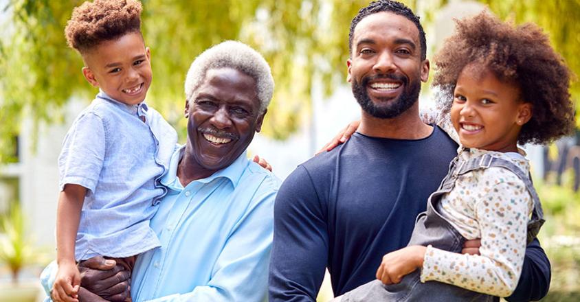 A bright daytime photo shows a smiling family of four together in a grassy yard. A man with a beard and a curly-haired child are smiling at the camera. The man is holding the child. Next to them is a smiling senior man holding another smiling curly-haired child. They are all wearing casual clothing. A house is visible in the background with greenery. 