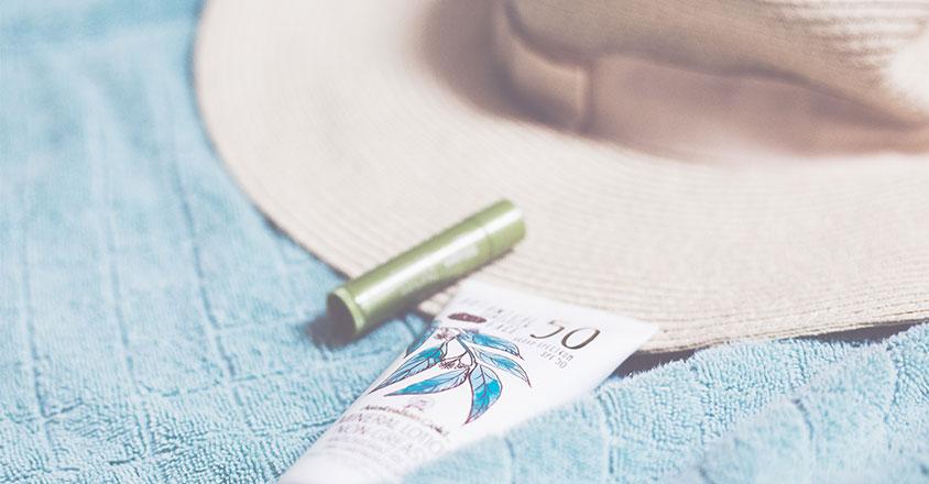 A tube of sunscreen with a leafy design, a green lip balm, and a wide-brimmed straw hat are neatly arranged on a textured, light blue towel, evoking thoughts of warmth and outdoor activities. 