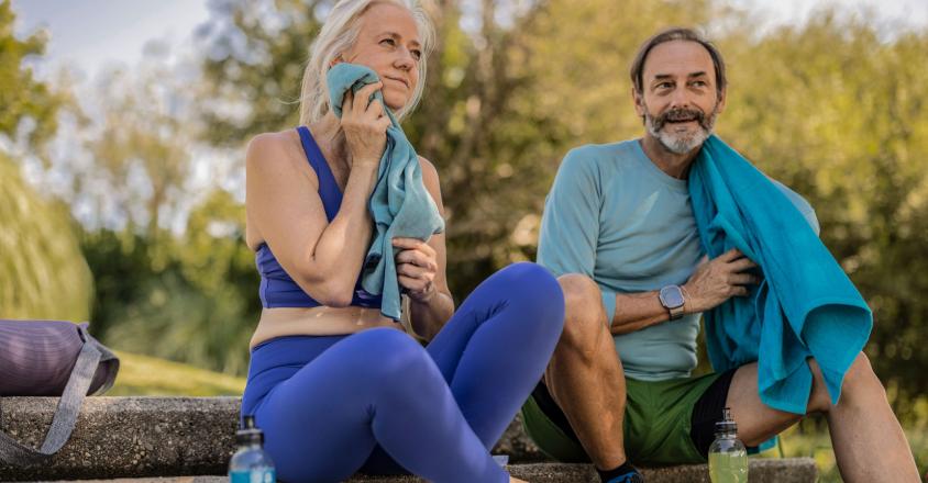 A woman with white hair and a man with a beard relax on stone steps outdoors after exercising. The woman wears a blue top and leggings and dries her face with a blue towel. The man wears a light blue long-sleeved shirt with green shorts, and a blue towel is draped over his shoulder.