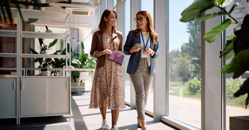 Two women stroll through a light-filled office hallway, engaged in a cheerful conversation. One woman, with light brown hair, wears a floral dress with a tan jacket, carrying a purple notebook. The other, with red hair and glasses, is dressed in a blue blazer, patterned pants, and is holding a mobile phone.
