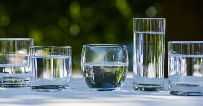 This outdoor shot features five glasses filled with clear water, placed on a white surface. The arrangement includes glasses of different shapes and sizes, creating a visually interesting display against a blurred green leafy backdrop. The lighting is bright and natural, emphasizing the clarity and purity of the water, and casting soft shadows on the surface. 