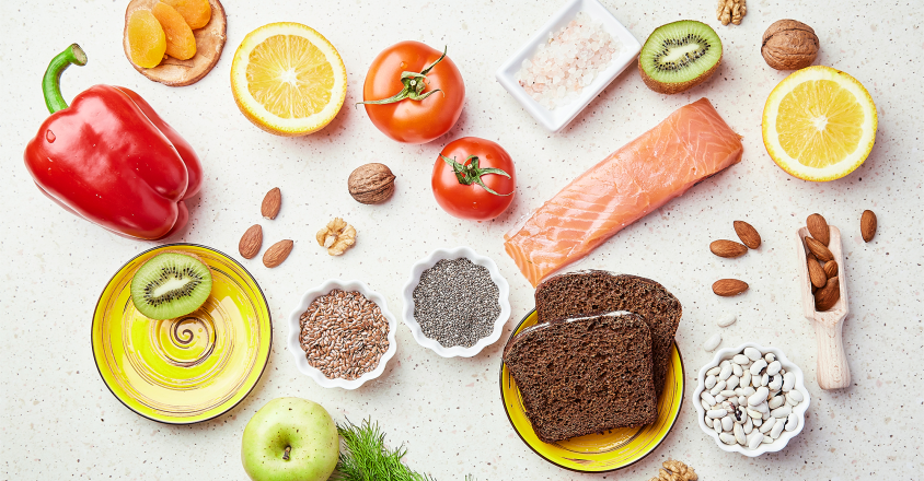Overhead shot of healthy fruits, vegetables, nuts, seeds and grains arranged on a white surface. A variety of colorful produce creates an appealing flat lay.