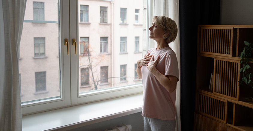 A mature woman with short blonde hair stands in a softly lit room near a large window, hands gently placed on her chest. She wears a pink t-shirt and grey pants.