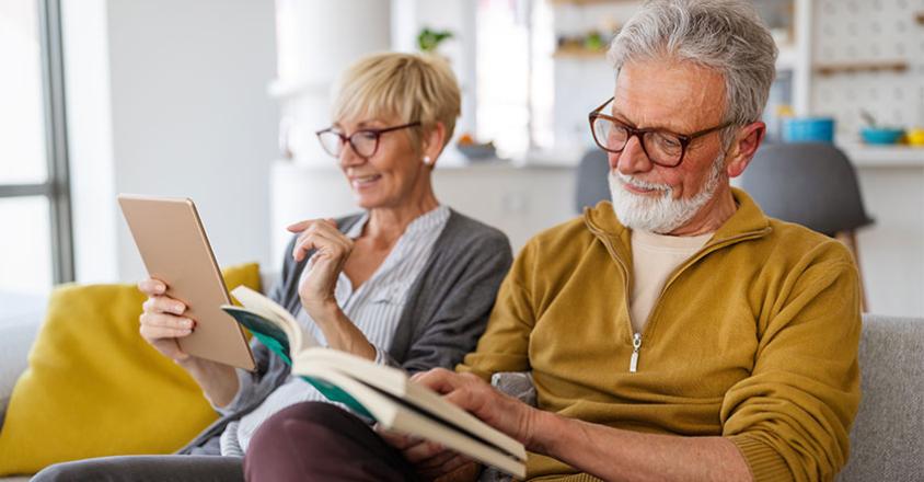 A mature couple relaxes at home in a brightly lit room. The woman with short blonde hair holds a tablet and smiles. A man with grey hair and a beard sits next to her and reads a book. 
