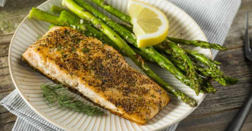 This close-up shot features a delectable grilled salmon fillet, served alongside fresh green asparagus spears and a vibrant lemon wedge. The salmon is seasoned and has a visually appealing texture. 