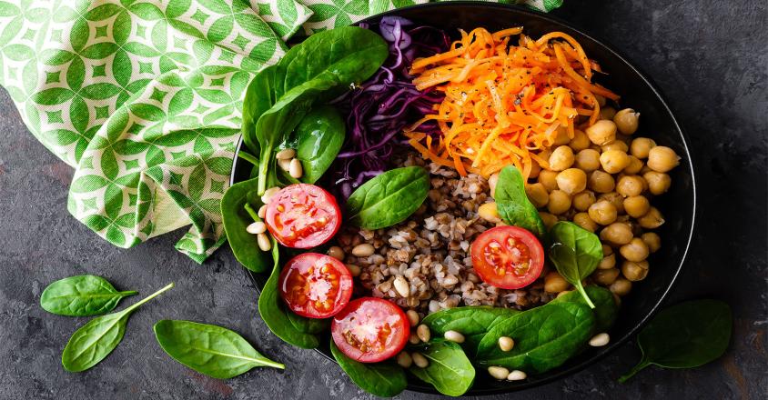 A vibrant vegan bowl is pictured from directly above, containing an assortment of fresh vegetables and grains. There is shredded orange carrots, red cabbage, chickpeas, spinach, tomatoes, grains and pine nuts in a black bowl.