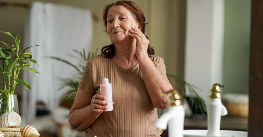 A senior woman with brown hair stands in a bathroom, holding a bottle of pink lotion. She is in front of a mirror, and has applied lotion to her face. The bathroom has neutral decor, with a white sink and leafy green plants. 