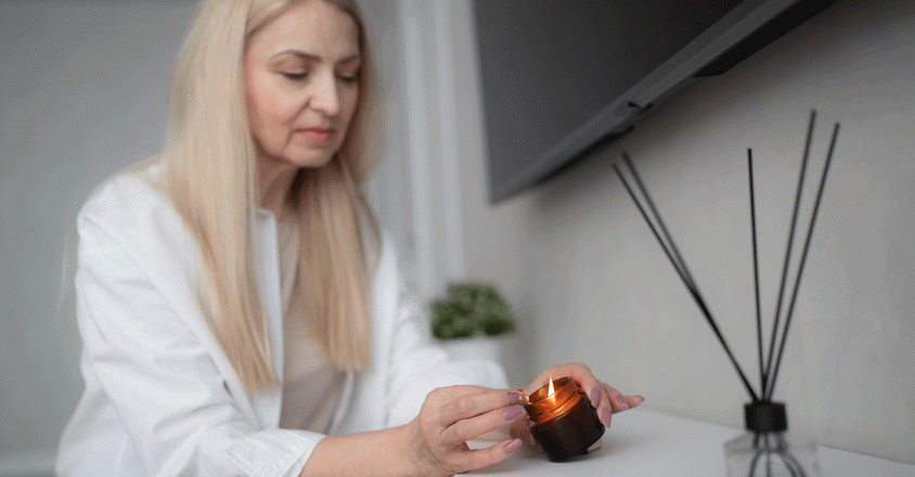 An adult woman with long blonde hair is lighting a small, amber-colored candle. The setting is indoors, possibly a home, with a white surface holding the candle and an aroma diffuser with black reeds. Above is a dark, flat screen television mounted on the wall. The soft lighting contributes to a calm mood.