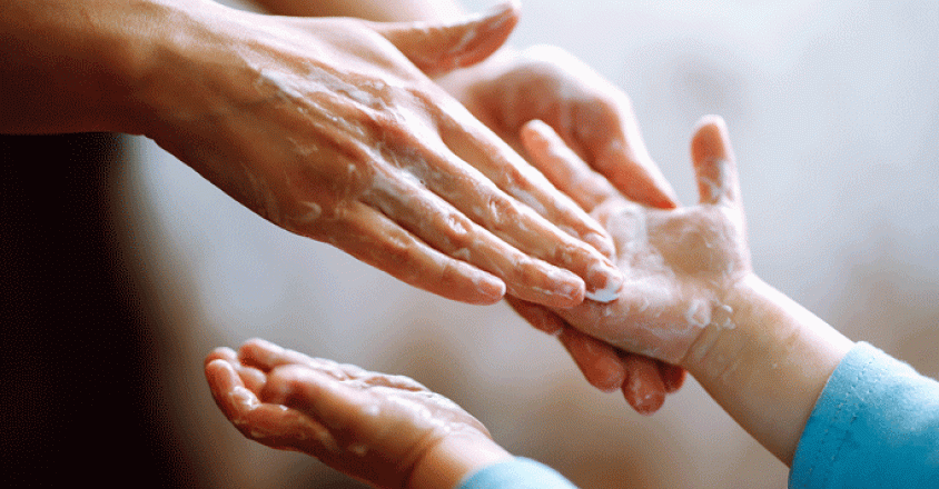 A close-up shot captures an adult carefully applying soap onto a child's hands. The hands are wet and covered in white soap.