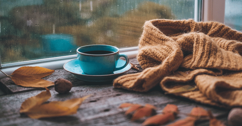 A cup of coffee sits by a window on a rainy day, complemented by a warm, knitted scarf and fallen leaves scattered on the wooden table. The tranquil scene evokes a sense of comfort and relaxation, ideal for projects centered on cozy moments, seasonal changes, or comforting beverages.