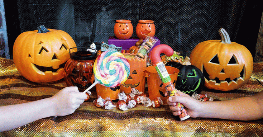 Two hands reach out to take Halloween candy. Several orange jack-o'-lantern pumpkins with scary faces are arranged with containers and wrapped candies. The shot is taken in a home. The background is a dark brick fireplace. The light in the photo is bright and even.