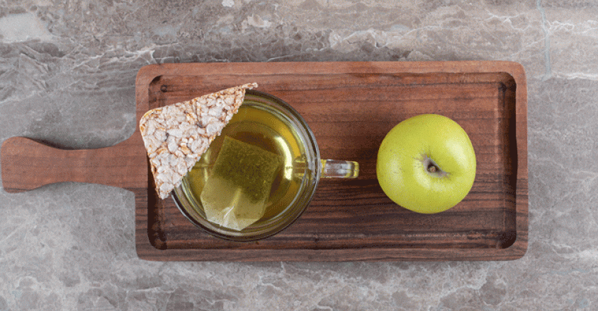 A top-down, bright shot depicts a clear mug of tea with a tea bag steeping, a triangular rice cake, and a whole green apple on a dark brown wooden tray. The tray sits on a grey and white mottled surface.