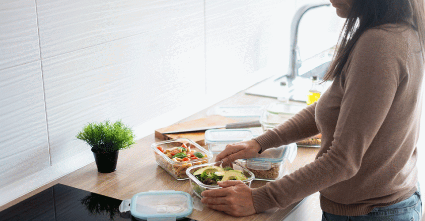A woman wearing a brown sweater and blue jeans stands in a bright, modern kitchen, organizing fresh meals into clear glass storage containers. The wooden countertop holds several prepared dishes, including a vibrant green salad topped with sliced avocado and a pasta salad with cherry tomatoes and herbs. 