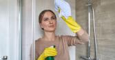 woman cleaning a glass shower with yellow gloves, spray bottle and towel