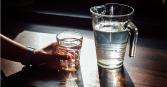 zoomed in perspective of a hand holding a clear glass of water with a large water pitcher nearby, against a dark backdrop and wood countertop 