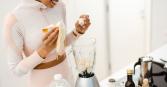 young woman in athletic wear smiles while adding a banana to a blender full of banana chunks on a white countertop. A bottle of milk, and various other food items sit on the counter.