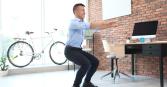 In a bright, naturally lit office, an adult man with short dark hair wearing a light blue button-down shirt, dark pants, and black shoes performs squats in front of a desk, computer, and brick wall. A bicycle stands near a window. The setting and subject suggests the concepts of active workplace, balance, and healthy habits. 