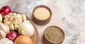 A wooden board holds a collection of culinary delights: red and white onions, fragrant garlic bulbs, and two wooden bowls filled with spices. The photo is shot in bright daylight, with a focus on the natural textures and colors of the ingredients, creating an inviting and wholesome mood. 
