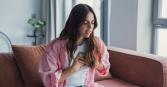 A young woman with wavy brown hair sits on a couch in a home interior. She wears a pink striped shirt, white t-shirt, and light blue jeans. She clutches her chest with both hands and appears to be in distress. The scene is brightly lit by daylight streaming in from nearby windows