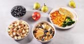 Eye-level shot presents a variety of healthy food bowls. A bowl of blueberries sits beside a bowl of sliced radishes and cucumbers. A bowl of cooked grain, apples, and blueberries is nestled near an apple and a couple of pears. A final bowl with cooked chicken, squash, and broccoli completes the spread.