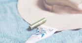 A tube of sunscreen with a leafy design, a green lip balm, and a wide-brimmed straw hat are neatly arranged on a textured, light blue towel, evoking thoughts of warmth and outdoor activities. 