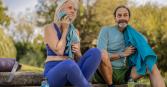 A woman with white hair and a man with a beard relax on stone steps outdoors after exercising. The woman wears a blue top and leggings and dries her face with a blue towel. The man wears a light blue long-sleeved shirt with green shorts, and a blue towel is draped over his shoulder.