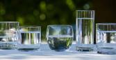 This outdoor shot features five glasses filled with clear water, placed on a white surface. The arrangement includes glasses of different shapes and sizes, creating a visually interesting display against a blurred green leafy backdrop. The lighting is bright and natural, emphasizing the clarity and purity of the water, and casting soft shadows on the surface. 