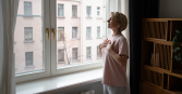 A mature woman with short blonde hair stands in a softly lit room near a large window, hands gently placed on her chest. She wears a pink t-shirt and grey pants.