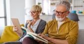 A mature couple relaxes at home in a brightly lit room. The woman with short blonde hair holds a tablet and smiles. A man with grey hair and a beard sits next to her and reads a book. 