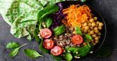 A vibrant vegan bowl is pictured from directly above, containing an assortment of fresh vegetables and grains. There is shredded orange carrots, red cabbage, chickpeas, spinach, tomatoes, grains and pine nuts in a black bowl.