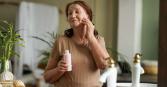 A senior woman with brown hair stands in a bathroom, holding a bottle of pink lotion. She is in front of a mirror, and has applied lotion to her face. The bathroom has neutral decor, with a white sink and leafy green plants. 