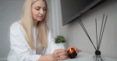 An adult woman with long blonde hair is lighting a small, amber-colored candle. The setting is indoors, possibly a home, with a white surface holding the candle and an aroma diffuser with black reeds. Above is a dark, flat screen television mounted on the wall. The soft lighting contributes to a calm mood.