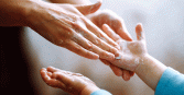 A close-up shot captures an adult carefully applying soap onto a child's hands. The hands are wet and covered in white soap.