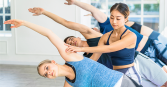 A group of adults practices yoga together indoors. Two women and two men in athletic attire are stretching in a bright studio with white walls and sunlight streaming in from windows. A woman is helping a blonde woman with a yoga pose. 