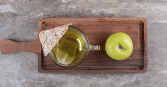 A top-down, bright shot depicts a clear mug of tea with a tea bag steeping, a triangular rice cake, and a whole green apple on a dark brown wooden tray. The tray sits on a grey and white mottled surface.