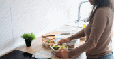 A woman wearing a brown sweater and blue jeans stands in a bright, modern kitchen, organizing fresh meals into clear glass storage containers. The wooden countertop holds several prepared dishes, including a vibrant green salad topped with sliced avocado and a pasta salad with cherry tomatoes and herbs. 
