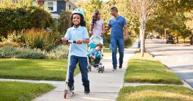 family of four taking a walk around the block with toddler wearing helmet and riding a scooter and baby in the stroller, mom and dad happy smiling
