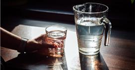 zoomed in perspective of a hand holding a clear glass of water with a large water pitcher nearby, against a dark backdrop and wood countertop 