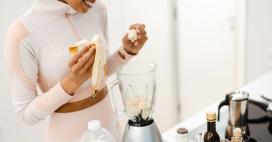 young woman in athletic wear smiles while adding a banana to a blender full of banana chunks on a white countertop. A bottle of milk, and various other food items sit on the counter.