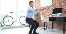 In a bright, naturally lit office, an adult man with short dark hair wearing a light blue button-down shirt, dark pants, and black shoes performs squats in front of a desk, computer, and brick wall. A bicycle stands near a window. The setting and subject suggests the concepts of active workplace, balance, and healthy habits. 