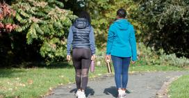 Two adults are walking on a paved path surrounded by trees. The women are wearing athletic clothing and carrying water bottles. The setting appears to be outdoors in a park-like environment with natural light.